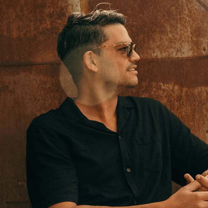 Man wearing sunglasses and a black shirt against a textured wall.
