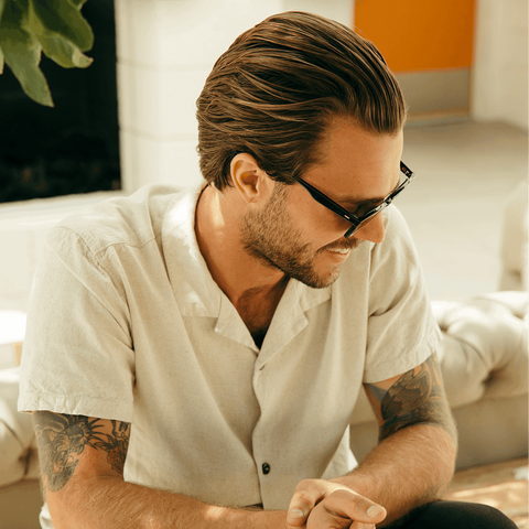 Man with tattoos wearing glasses and a white shirt sitting on a couch.