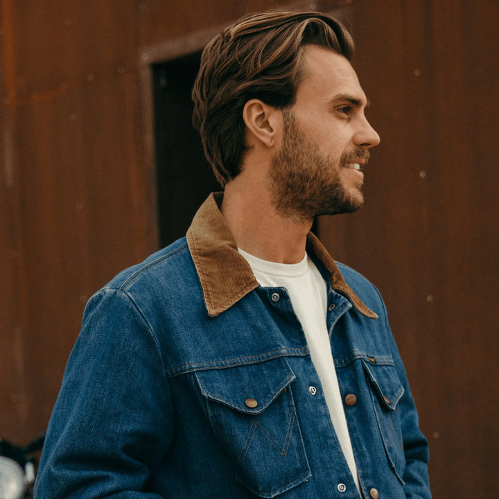 Man wearing a blue denim jacket with a brown collar against a rust-colored wall.